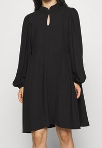 Svart klänning med nyckelhålsringning, långa ärmar och samlad midja. Tillverkad av ett mjukt material har den en flowy silhuett och knälång fåll.