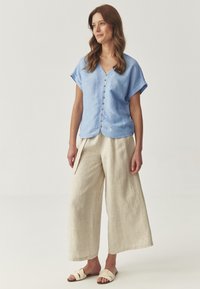 Lichtblauw linnen top met korte mouwen en knopen, gecombineerd met losse beige linnen broek en lichtgekleurde sandalen. Eenvoudig ontwerp en textuur.