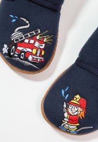 Chaussons en tissu bleu marine avec semelles en caoutchouc ornés de motifs brodés colorés représentant un camion de pompiers et un pompier.