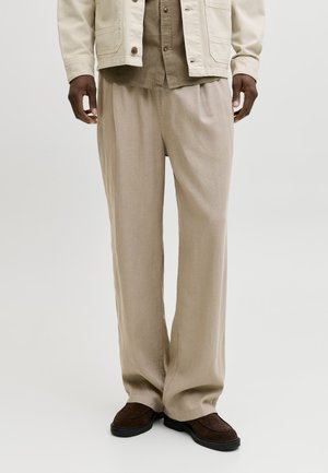 Férfi bézs színű, bőszárú nadrágban, barna cipőben, olívazöld ingben és törtfehér zakóban, egyszínű háttér előtt.