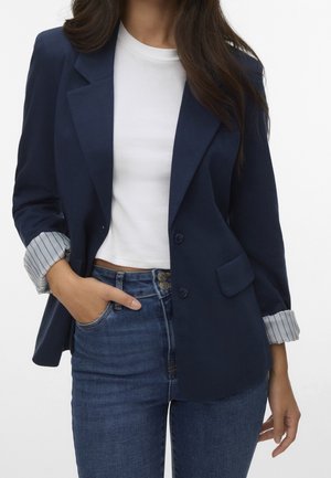 Woman wearing a navy blazer with striped cuff lining over a white crop top and blue high-waisted jeans, one hand in pocket.