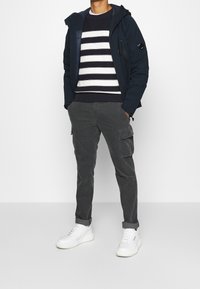 Granatowa kurtka z kapturem, biało-granatowy sweter w paski, szare spodnie cargo i białe sneakersy. Posiada zapięcia na suwak oraz naszywki na rękawach.