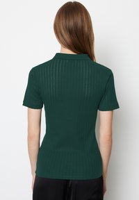 Kobieta z długimi brązowymi włosami, ubrana w ciemnozielony, prążkowany, krótkorękawowy sweter, pokazana z tyłu na białym tle.