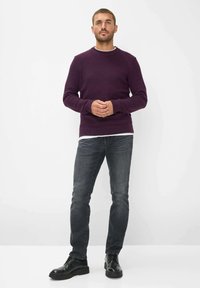 Donkerpaarse trui met geribde boorden en zoom, gedragen over een wit shirt. Gecombineerd met grijze spijkerbroek en zwarte veterschoenen. Eenvoudig, aansluitend ontwerp.