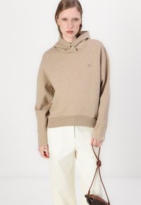 Beige hoodie met een relaxed fit en een trekkoordcapuchon. Rib gebreide zoom en boorden, zachte textuur. Gecombineerd met crèmekleurige broeken en een bruine tas.