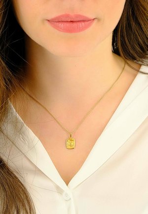 Collier en pendentif en or avec un charme rectangulaire, présentant des détails complexes, suspendu à une fine chaîne, porté sur un chemisier blanc.