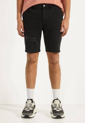 Mann trägt schwarze zerrissene Jeansshorts, weiße Sportsocken und graue-weiße klobige Sneakers, steht vor weißem Hintergrund.