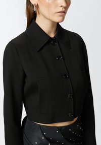 Kratka crna jakna sa strukturiranom kragnom, prednjim dugmadima i dugim rukavima; sadrži krojene šavove i glatku teksturu.