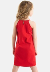 Niña joven con largo cabello ondulado, de pie de espaldas, vistiendo un vestido rojo sin mangas con un pequeño detalle de botón en forma de llave en la parte de atrás.