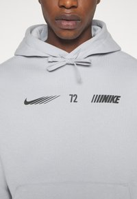 Homme portant un sweat à capuche gris clair avec des logos Nike et le numéro 72 imprimé en noir sur la poitrine, vue de face recadrée au niveau de la bouche.