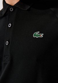 Černé polo tričko s klasickým límcem, třemi knoflíky a zeleným krokodýlím logem s červenými detaily na levé části hrudi.