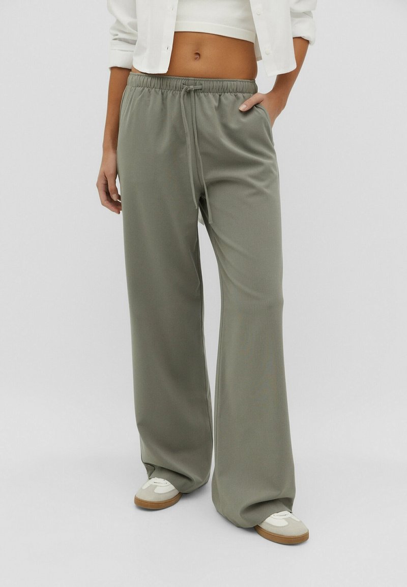 Personne portant un pantalon ample vert olive avec cordon, un crop top blanc, une veste blanche et des baskets blanches à semelles en caoutchouc.