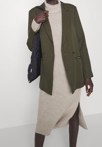 Blazer verde oliva con bottoni doppi, indossato sopra un abito lavorato a maglia beige con uno spacco laterale e orlo a coste. Borsa nera appoggiata sulla spalla.