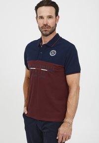Polo marškiniai su tamsiai mėlynais ir burgundiškais spalvų blokais, trumpomis rankovėmis, apykakle ir apskritu logotipu kairėje pusėje. Priekyje atspausdintas tekstas.