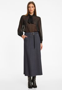 Frau trägt eine transparente schwarze Bluse mit langen Ärmeln und Krawatte, einen grauen Midirock mit Nadelstreifen und Gürtel sowie schwarze Stiefel, steht mit einer Hand in der Tasche.