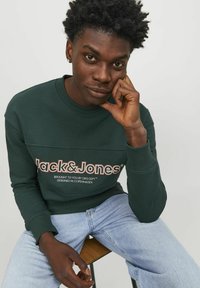 Sudadera verde oscuro con escote redondo y puños acanalados, que presenta texto de logotipo en blanco y beige. Jeans azul claro de corte relajado.