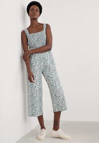 Monos de estampado floral con cuerpo fruncido, piernas anchas y cortas, y tirantes ajustables en azul verdoso y blanco. Combinado con zapatillas beige.