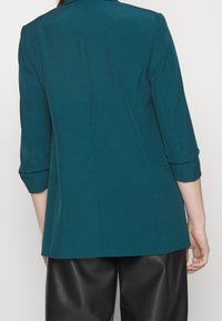 Turkusowa, dopasowana marynarka z wyciętym kołnierzem, rękawami o długości trzech ćwiartek oraz gładką teksturą materiału. Charakteryzuje się czystym, dopasowanym krojem z tyłu.