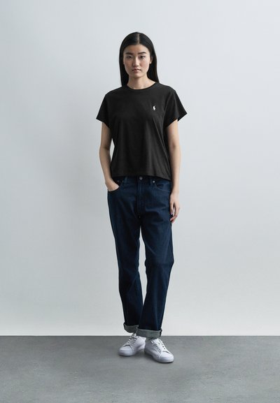 Νεαρή γυναίκα στέκεται μπροστά σε μονόχρωμο γκρι τοίχο, φορώντας μαύρο κοντομάνικο πουκάμισο, σκούρα μπλε τζιν με μανσέτες και λευκά αθλητικά παπούτσια, με τα χέρια στις τσέπες.