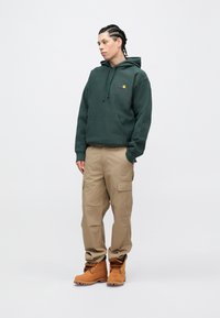 Sudadera con capucha verde oscuro con bolsillo frontal y logo rojo-amarillo, combinada con pantalones cargo beige y botas color canela con herrajes dorados.