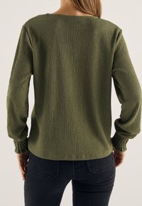 Top texturizado verde oliva con un ajuste holgado, mangas largas y puños fruncidos, combinado con jeans ajustados negros. Vista trasera mostrada.