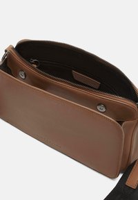 Braune Leder-Crossbody-Tasche mit offenem Hauptfach, innerer Reißverschlusstasche, magnetischem Klappenverschluss und teilweise sichtbarem schwarzen Stoffriemen.