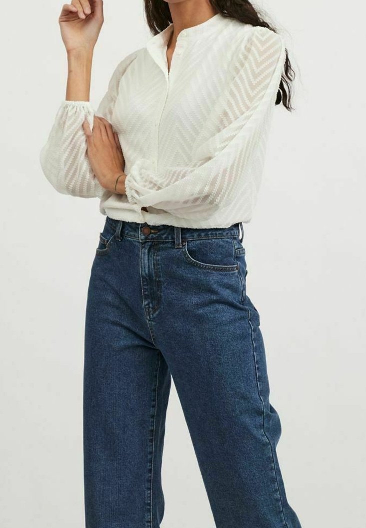 Valkoinen teksturoitu paita, jossa on soikio kuvio, korkea kaulus ja pallomaiset hihat, yhdistettynä sinisiin korkeavyötäröisiin farkkuihin, joissa on napitettu sulku.