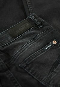 Närbild av svarta denimbyxor som visar bältesögglans, framfickan med metallnitar och en svart märkesetikett sydd på midjebandet.