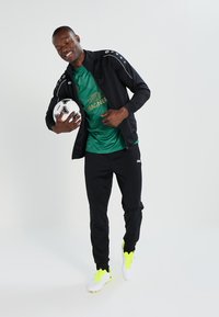 Survêtement noir avec détails blancs, t-shirt vert avec logo doré, tenant un ballon de football, et portant des chaussures de football jaunes vives.