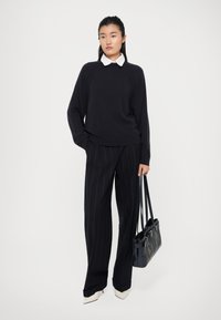 Maglione navy, camicia bianca con colletto, pantaloni a righe e tacchi color crema. Gli accessori includono una borsa nera con manici.