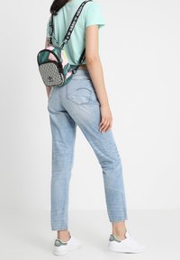 Mały plecak w kolorze czarnym i zielonym z geometrycznym wzorem, z różowymi akcentami i regulowanymi ramiączkami, noszony na jasnoniebieskiej bluzce.