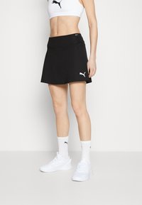 Falda atlética negra con una textura suave, que presenta una cintura alta y un pequeño logo de Puma blanco en el lado izquierdo. Complementada con zapatillas blancas.