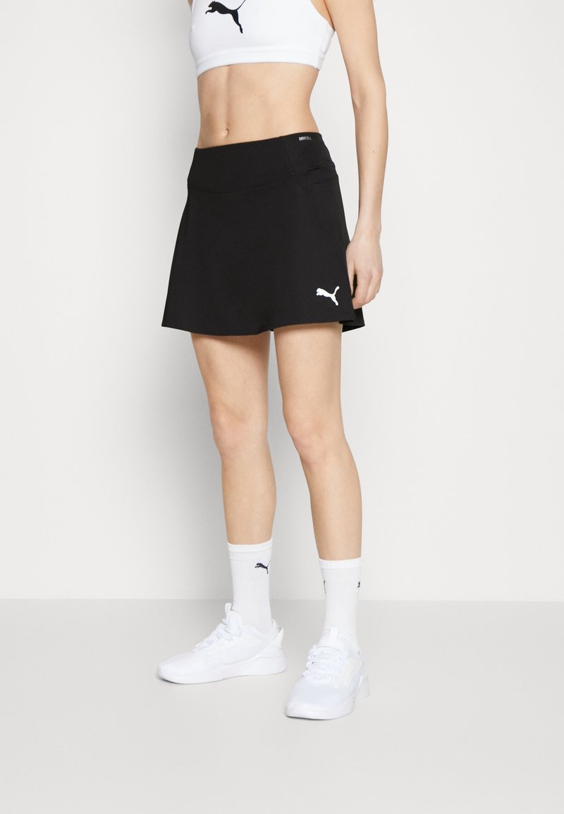 Falda atlética negra con una textura suave, que presenta una cintura alta y un pequeño logo de Puma blanco en el lado izquierdo. Complementada con zapatillas blancas.