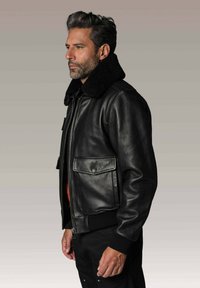 Homme aux cheveux et à la barbe grises portant une veste en cuir noir avec un grand col en fourrure, se tenant de profil sur un fond gris.