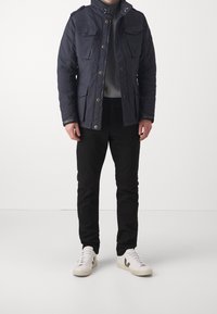 Granatowa kurtka wierzchnia z wieloma kieszeniami, zamkami błyskawicznymi i guzikami; w zestawieniu z czarnymi spodniami i beżowymi trampkami.