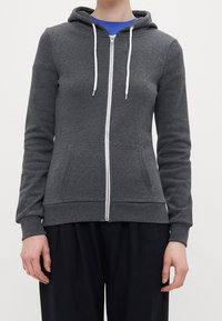 Sudadera con cremallera gris hecha de tela suave, con cierre frontal, cordones blancos y dos bolsillos frontales con puños y bajo de canalé.