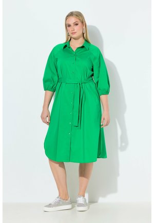 Žena v jasně zelených midi šatech na knoflíky s tříčtvrtečními rukávy a zavazováním v pase, doplněná třpytivými stříbrnými teniskami.