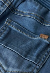 Zoom na denim tkanino, ki prikazuje majhno zadnjo žep s kontrastnim zlatim šivanjem in usnjenim etiketom z napisom "imej to". Prevladujejo modri odtenki.