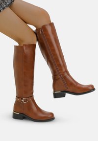 Botas marrones hasta la rodilla hechas de material sintético suave, con una cremallera lateral, una correa con hebilla y un tacón bajo tipo bloque.