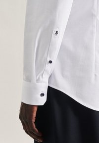 Dunkelhäutige Hand eines Mannes neben weißem Hemdsärmel mit schwarzen Knöpfen und kleiner schwarzer Rosenstickerei am Ärmelaufschlag.