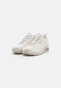 Baskets synthétiques blanches avec une texture lisse, un design à lacets et un talon légèrement surélevé. Présente une marque sur la languette et les panneaux latéraux.