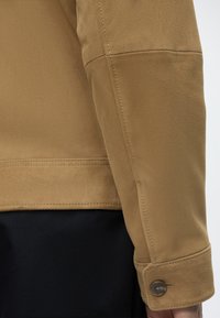 Le bras d’une personne dans une veste en daim beige avec un poignet boutonné, à côté d’un pantalon noir, vu de côté sur un fond clair.