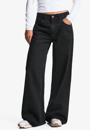 Personne portant un jean noir ample à jambes larges avec des baskets blanches et un top court blanc à manches longues, main dans la poche.
