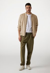 Beige bomberjack over een wit T-shirt, gecombineerd met olijfgroene broeken en witte sneakers. De jas heeft een ritsluiting en ribgebreide manchetten.
