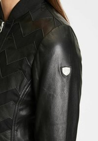 Svart läderbomberjacka med texturerat chevronmönster, ribbad krage och silverfärgad logobadge på vänster ärm. Slät yta.
