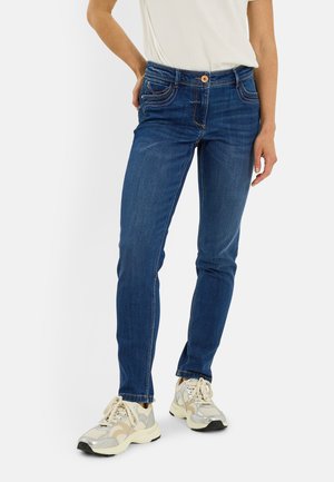 Personne portant un jean slim bleu et des baskets blanches, une main posée sur la hanche et un haut blanc rentré dans le pantalon.