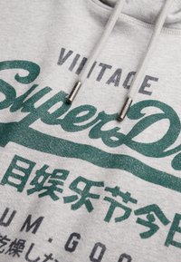 Felpa con cappuccio grigia con testo vintage verde e grigio "SuperDry." Caratterizzata da coulisse con punte in metallo. Tessuto morbido leggermente strutturato.