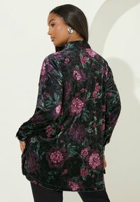 Camicia di velluto nera con un ampio motivo floreale viola. Presenta un colletto, maniche lunghe e spacchi laterali per facilitare i movimenti.