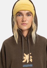 Brauner Hoodie mit einem großen gelb-blauen bestickten Blumenmuster und "Quiksilver"-Schriftzug. Getragen mit einer gelben Rippmütze.