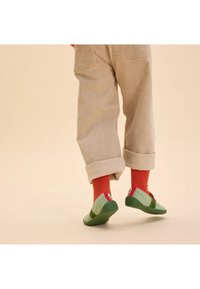 Zielono-szare buty wsuwane z teksturowanej tkaniny, z kontrastującymi czerwonymi akcentami i elastyczną gumową podeszwą, noszone z podwiniętymi beżowymi spodniami i pomarańczowymi skarpetkami.